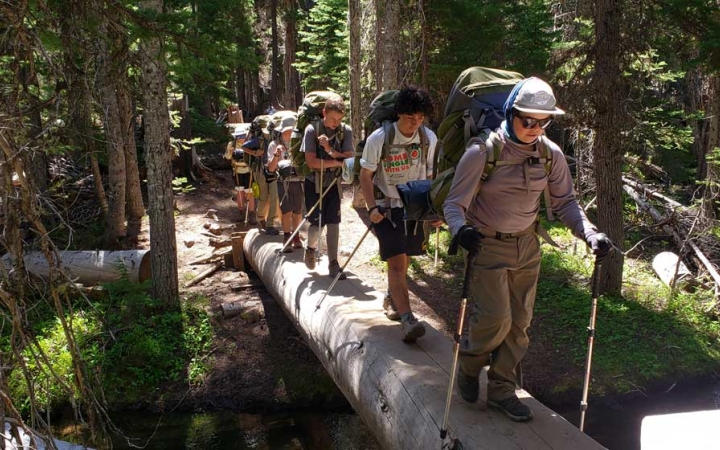 A group of teens wearing backpacks and using trekking poles hike over a wood plank